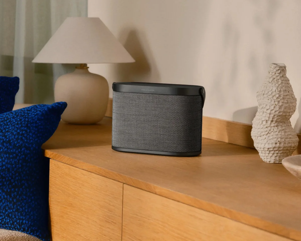 Bang & Olufsen portable wireless speaker in dark grey fabric, positioned on a wooden sideboard.