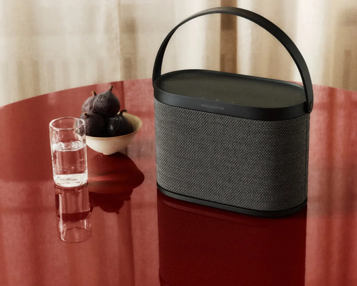 Bang & Olufsen portable wireless speaker featuring a dark grey fabric grille and a black carrying handle, positioned on a glossy red table beside a glass of water and a bowl of figs.