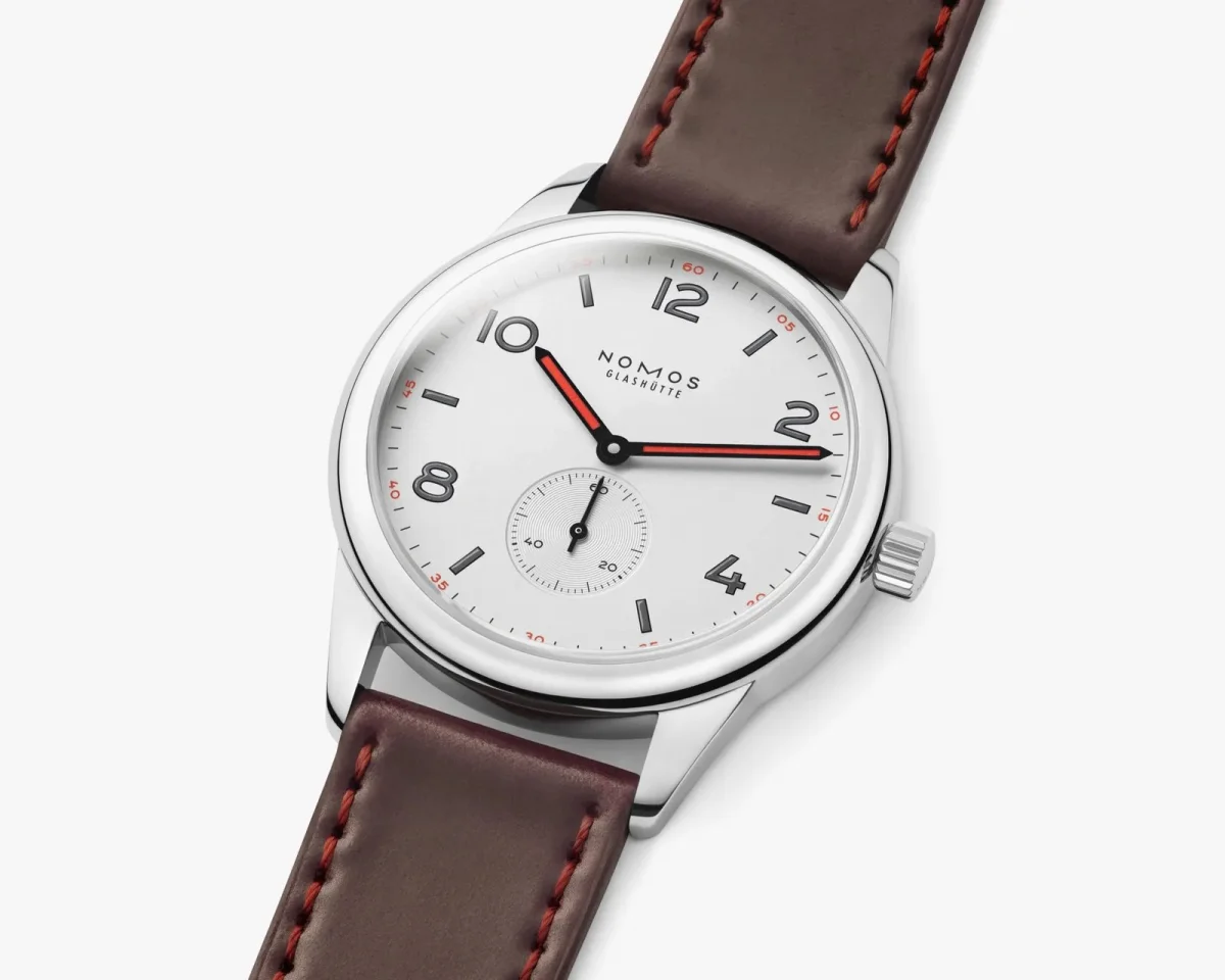 Nomos Glashütte wristwatch featuring a white dial with black numerals and red hands, paired with a brown leather strap accented by red stitching.