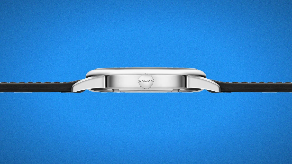 Side view of a silver Nomos watch case featuring a branded winding crown attached to a dark leather strap, set against a blue background.