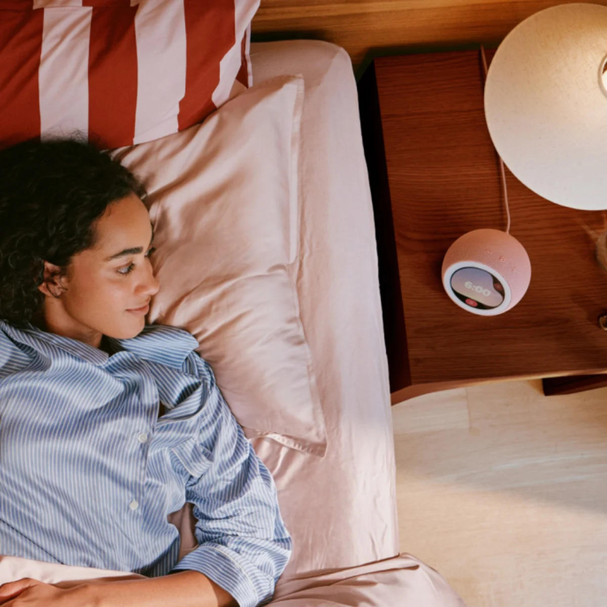 Round, pink smart speaker with a digital clock display on a nightstand.