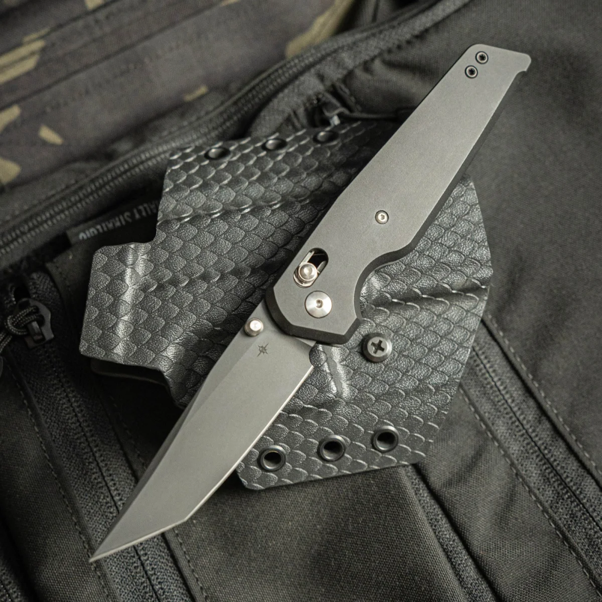 Grey folding knife with a tanto blade resting on a textured, dark grey holster.