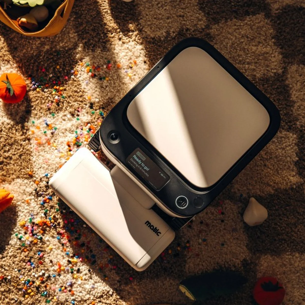 Matic robotic floor cleaner on a patterned rug near spilled colorful beads and toy vegetables.