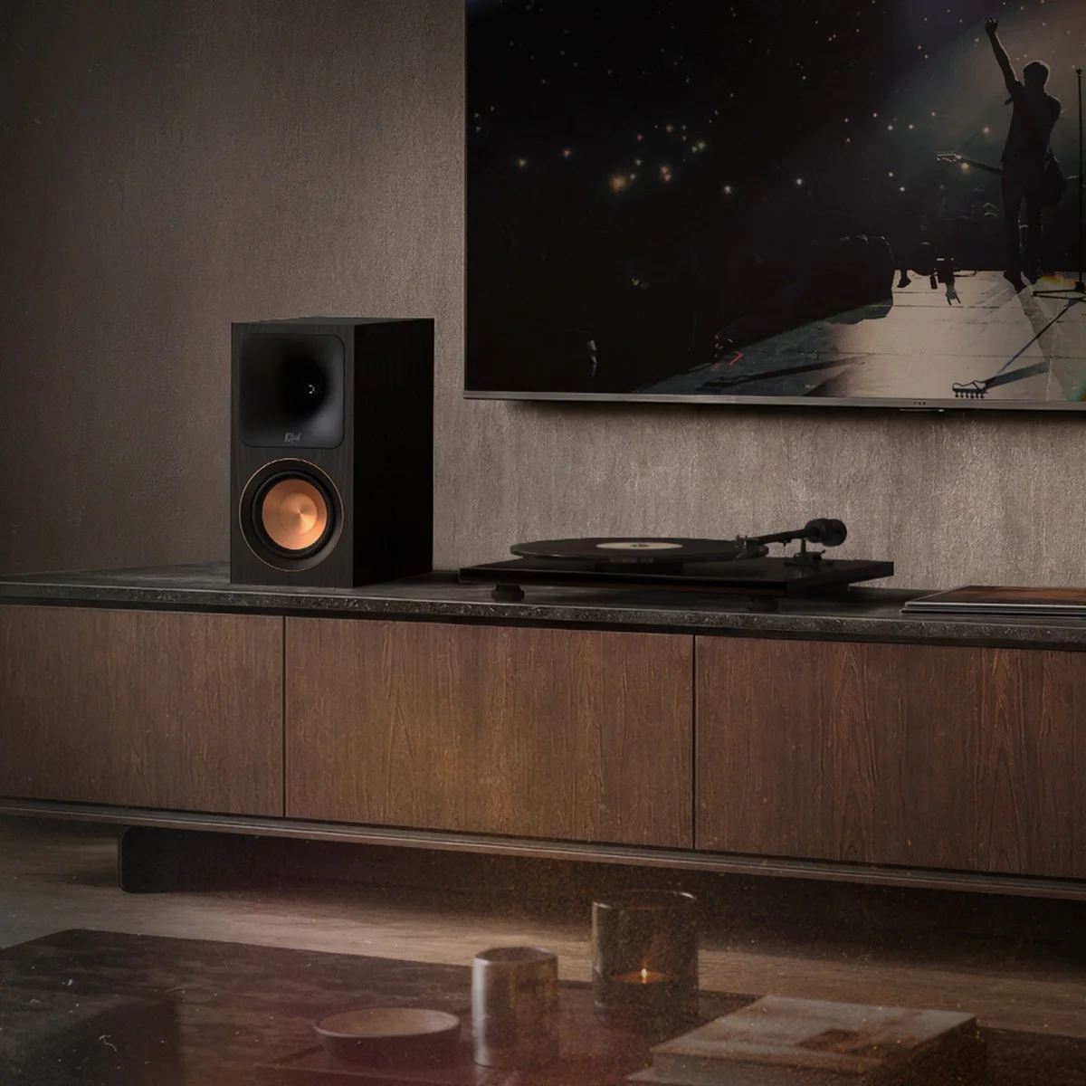 Black bookshelf speaker with copper cone woofer on a wooden cabinet under a wall-mounted TV.
