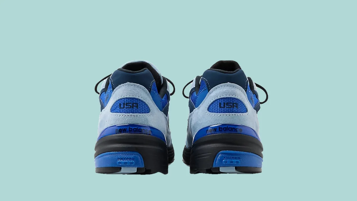 Blue New Balance sneakers shown from the back, with USA text on suede heels and black and blue soles, positioned against a light blue background.