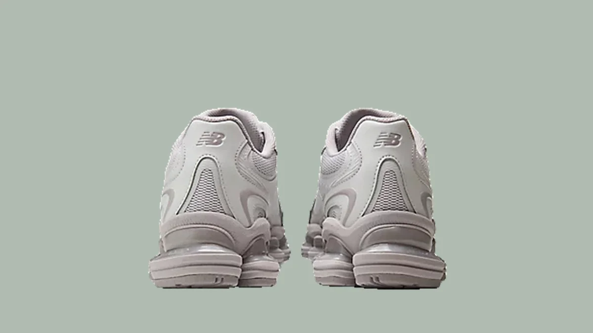 Light grey New Balance sneakers viewed from the back, featuring the NB logo on the heels, set against a plain light green background.