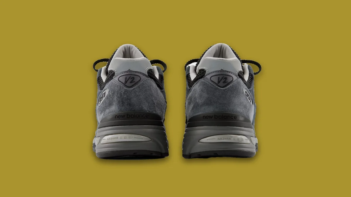 Rear view of two grey New Balance 991v2 sneakers placed on an olive green background.
