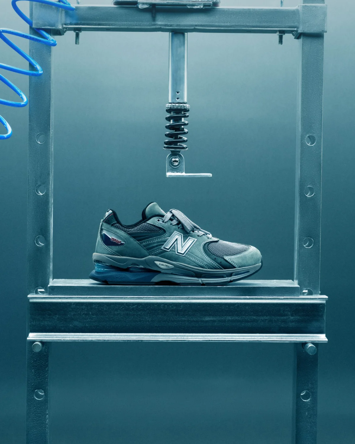 Gray New Balance sneaker with blue accents under a metal compression testing machine.