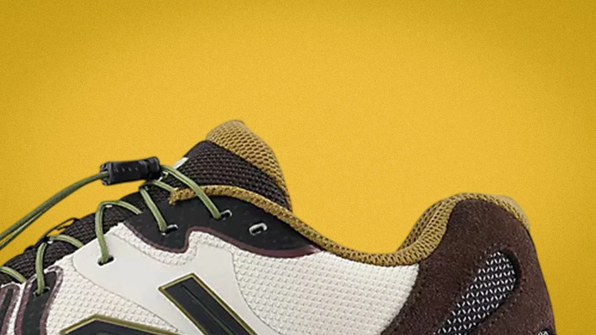 Close-up of a sneaker with beige mesh, black and brown suede, olive green laces, and a yellow background.