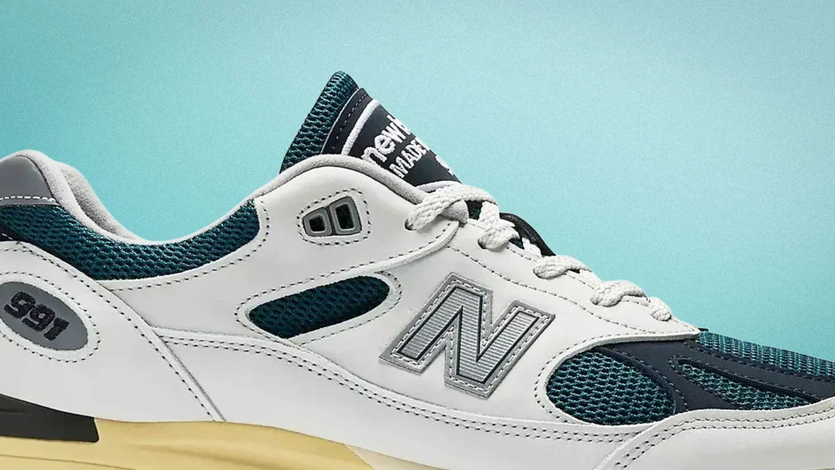 White and teal New Balance 991 sneaker with mesh panels and beige sole against a light blue background.