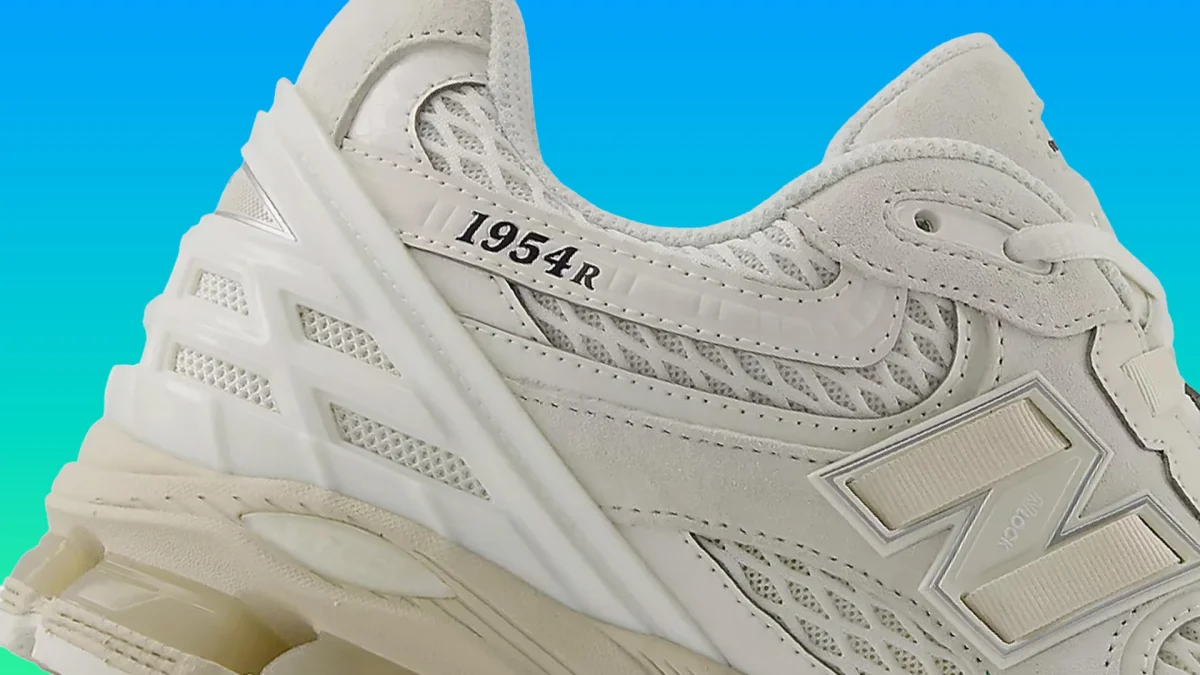 Close-up of a beige New Balance 1954R sneaker with mesh and suede details against a blue gradient background.