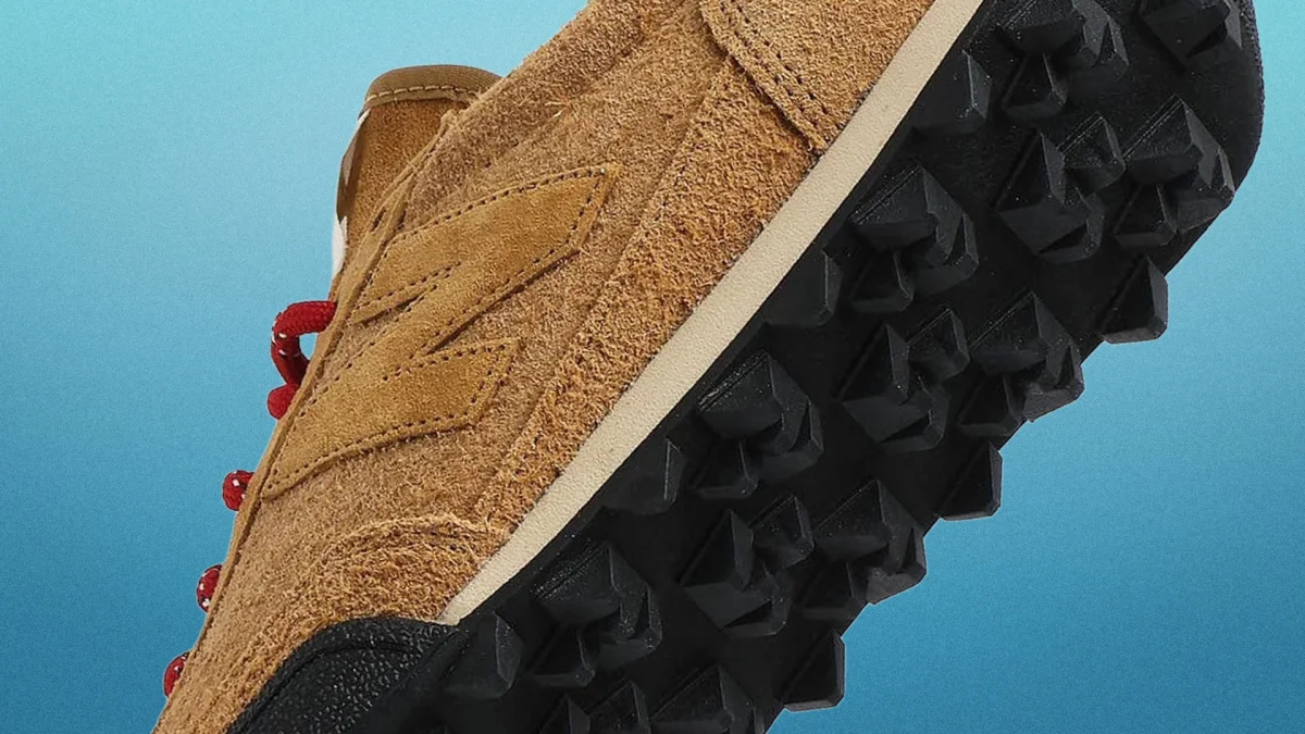 Brown suede sneaker with red laces and black studded sole against a blue gradient background.