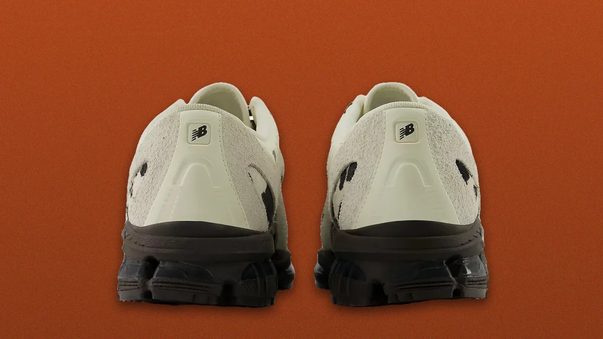 Two off-white New Balance sneakers shown from the back, featuring dark brown soles, textured off-white heels with the New Balance logo, and black detailing, set against a solid orange background.