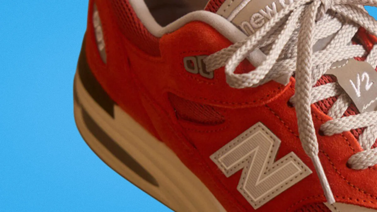 Red New Balance sneaker with white laces and beige midsole against a blue background.
