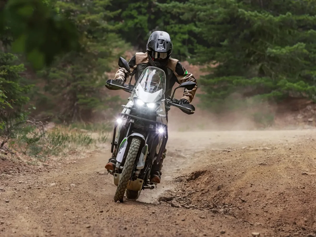 Motorcyclist in helmet and riding gear leaning into a turn on a dusty dirt road surrounded by trees.