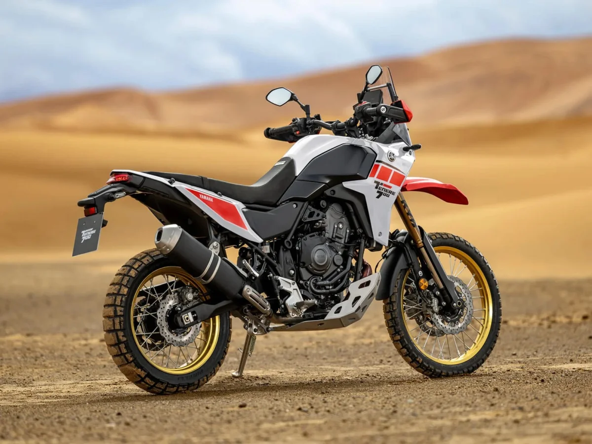 Yamaha Ténéré 700 dual-sport motorcycle in white with red graphics and gold rims, parked on a desert landscape with sand dunes in the background.