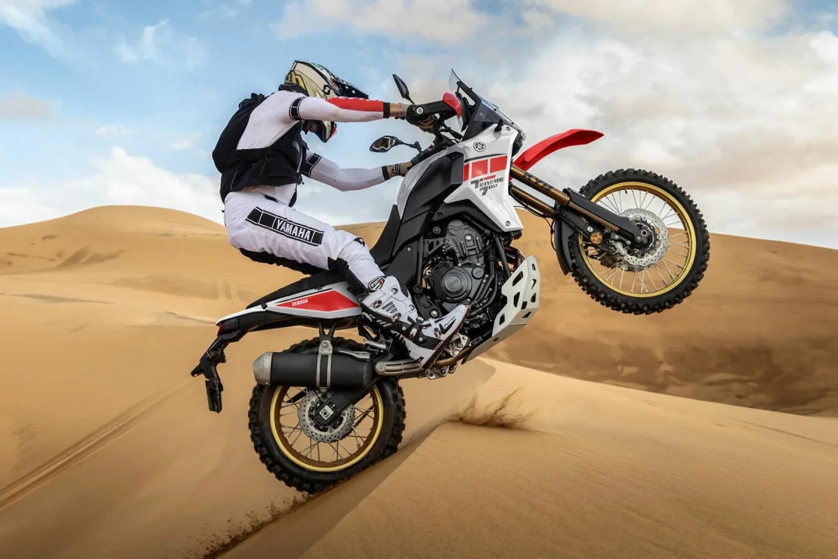 White and red Yamaha Ténéré 700 motorcycle performing a wheelie on a sand dune.