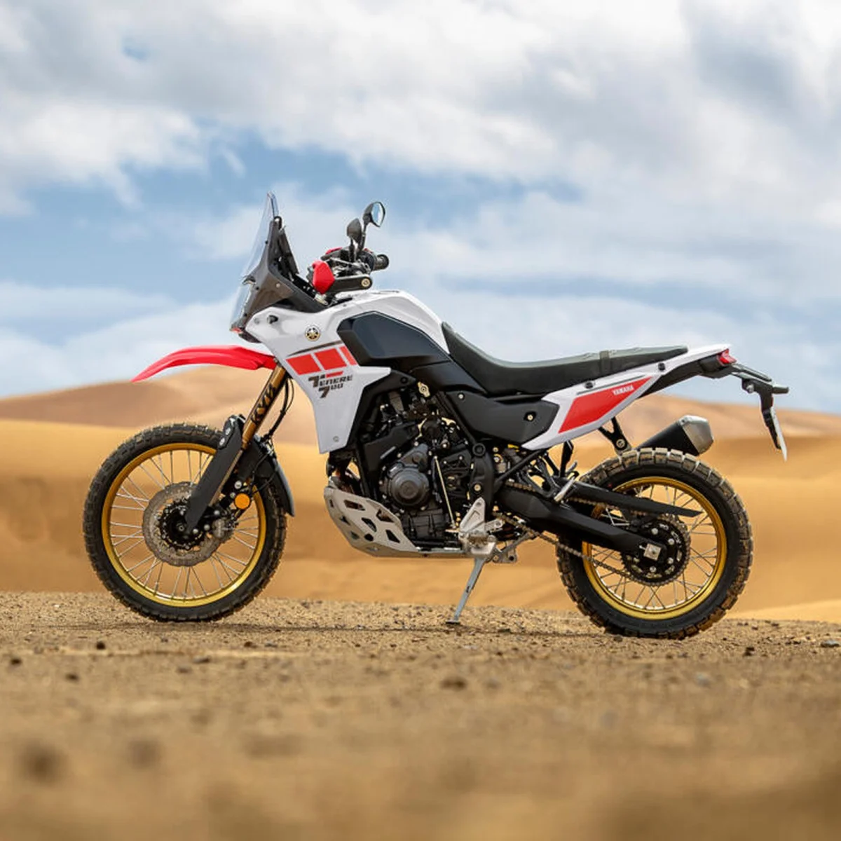 Yamaha Ténéré 700 adventure motorcycle with white and red bodywork, gold-rimmed spoked wheels, and a skid plate, parked in a sandy desert.