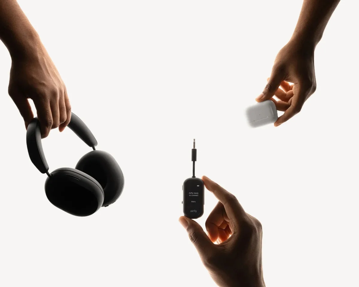 Black over-ear headphones, a white wireless charging case, and a small black AirFly wireless transmitter with a 3.5mm plug held in hands.