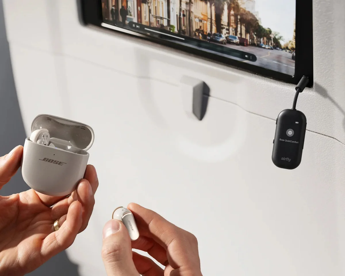 Hands holding a pair of Bose wireless earbuds and their open charging case, with a Twelve South AirFly Bluetooth transmitter plugged into an airplane seatback screen.