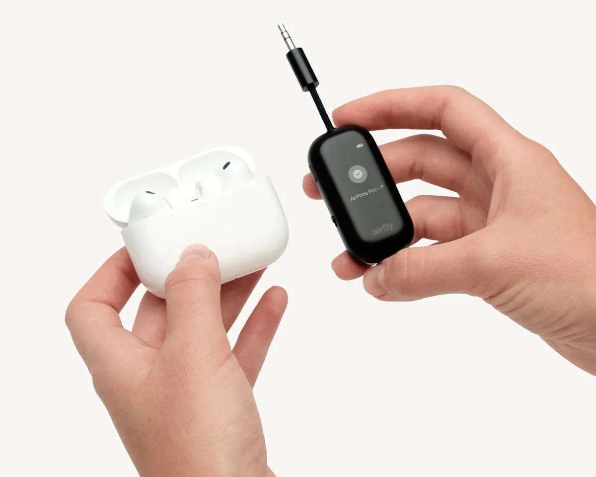 A white wireless earbud charging case and a black AirFly Bluetooth transmitter with a 3.5mm jack held in a hand.