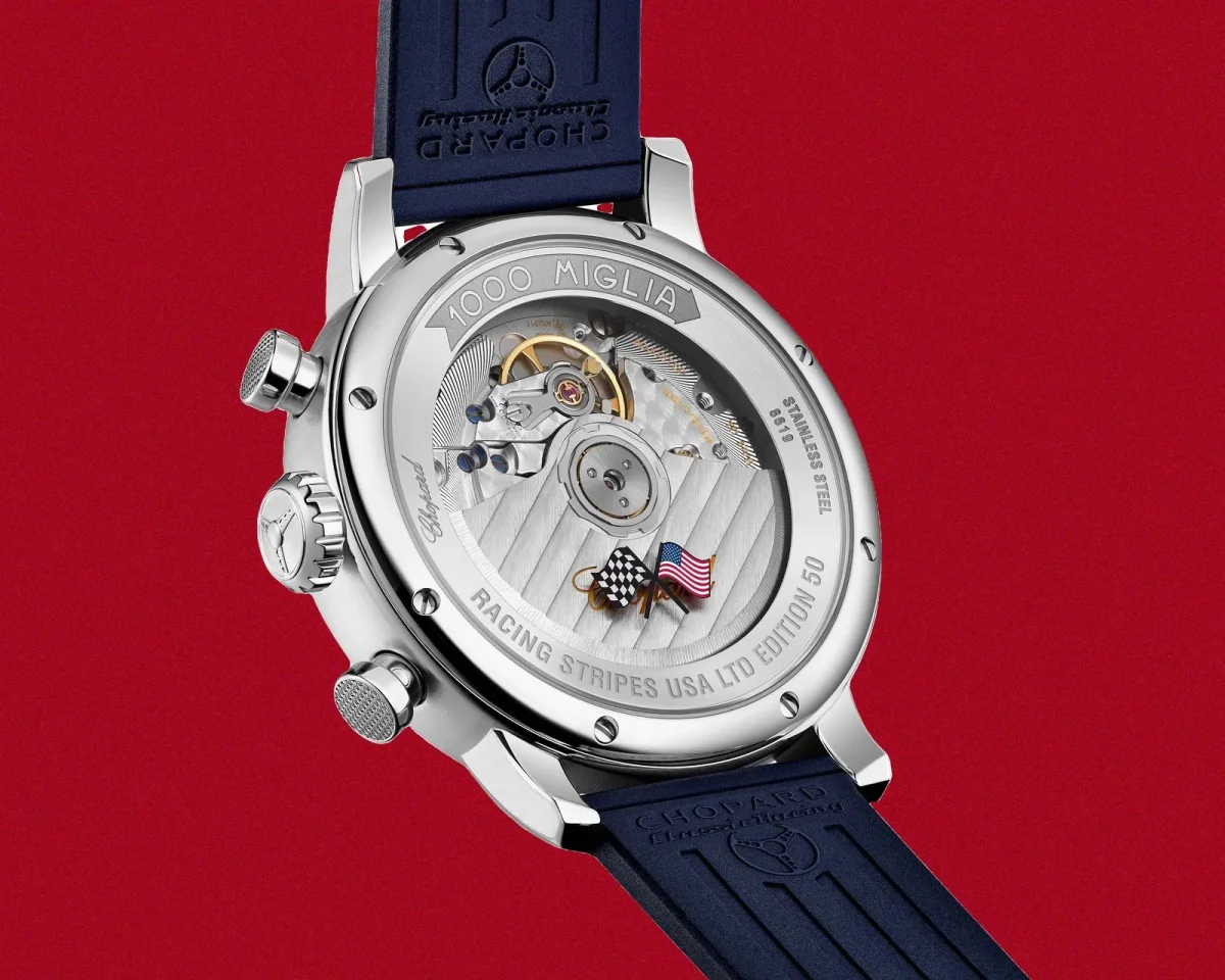 Back view of a stainless steel Chopard Mille Miglia watch featuring an exhibition case back with a visible mechanical movement, a checkered flag and US flag emblem, and a blue rubber strap, set against a red background.
