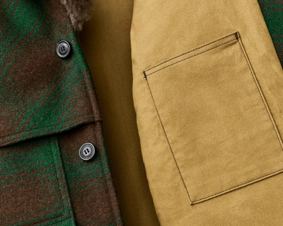 Close-up of a green and brown plaid wool coat featuring dark buttons and a tan interior lining with a square patch pocket.