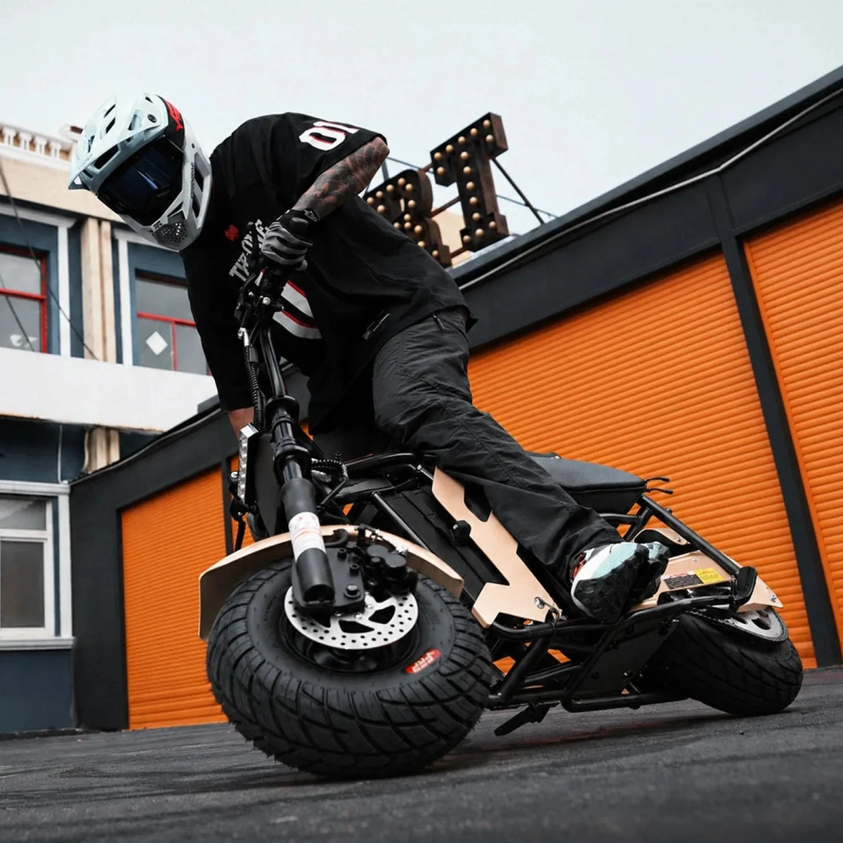 A tan-colored, wide-tire electric scooter is ridden by an individual wearing a full-face helmet in front of a building with orange garage doors.