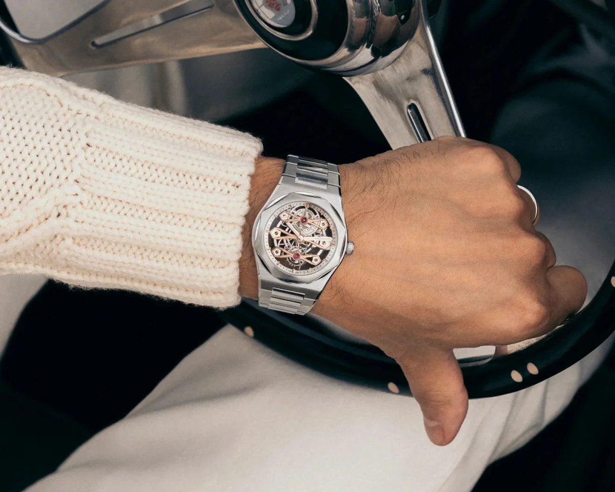 Silver skeleton watch with an exposed mechanical movement and stainless steel bracelet, worn on a wrist resting on a steering wheel.