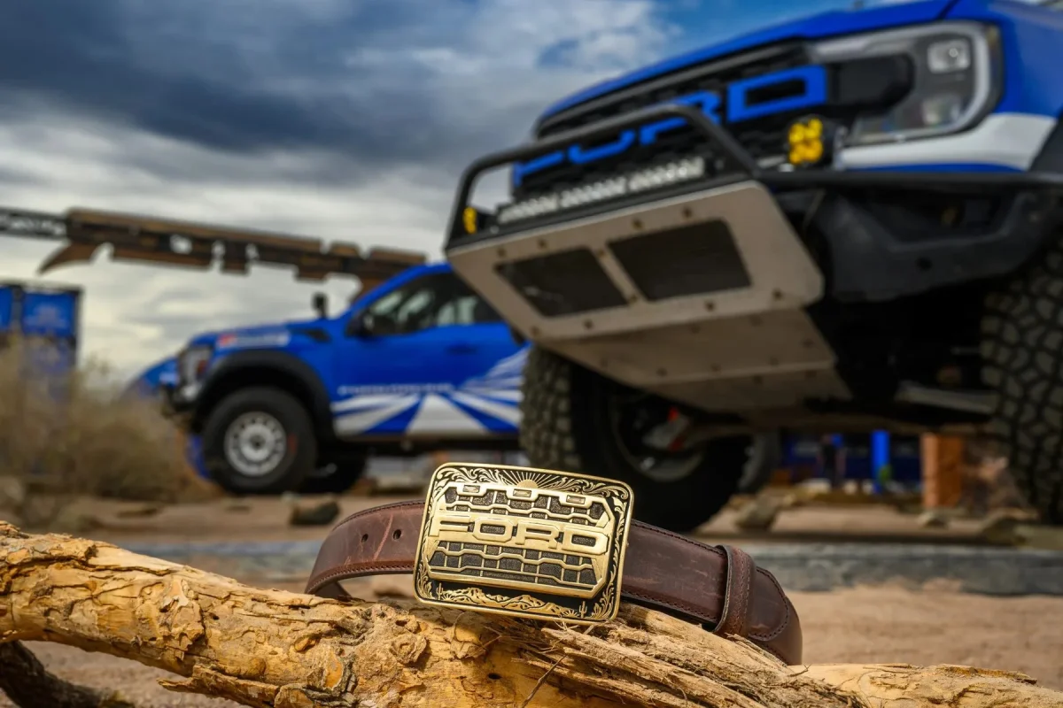 Brown leather belt with a rectangular brass buckle featuring a Ford truck grille design, resting on a log with two blue Ford trucks in the background.