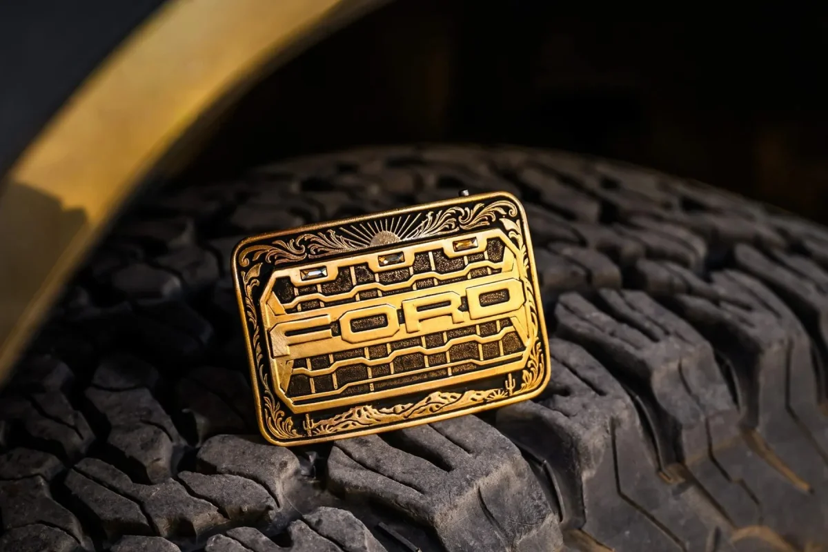 Gold-toned rectangular belt buckle featuring an engraved Ford logo and grille design, resting on a tire.