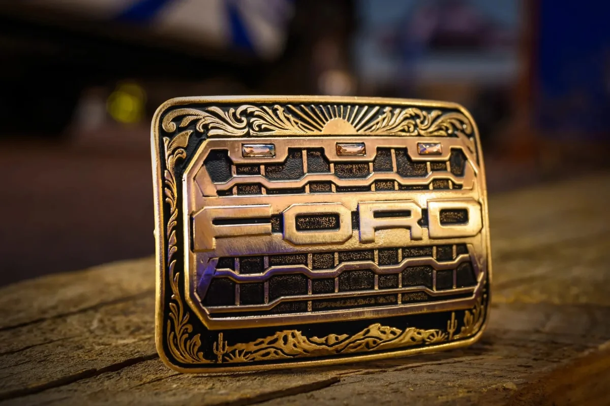 Rectangular belt buckle featuring the Ford logo set against a stylized grille pattern, framed by ornate engraved floral and desert motifs.