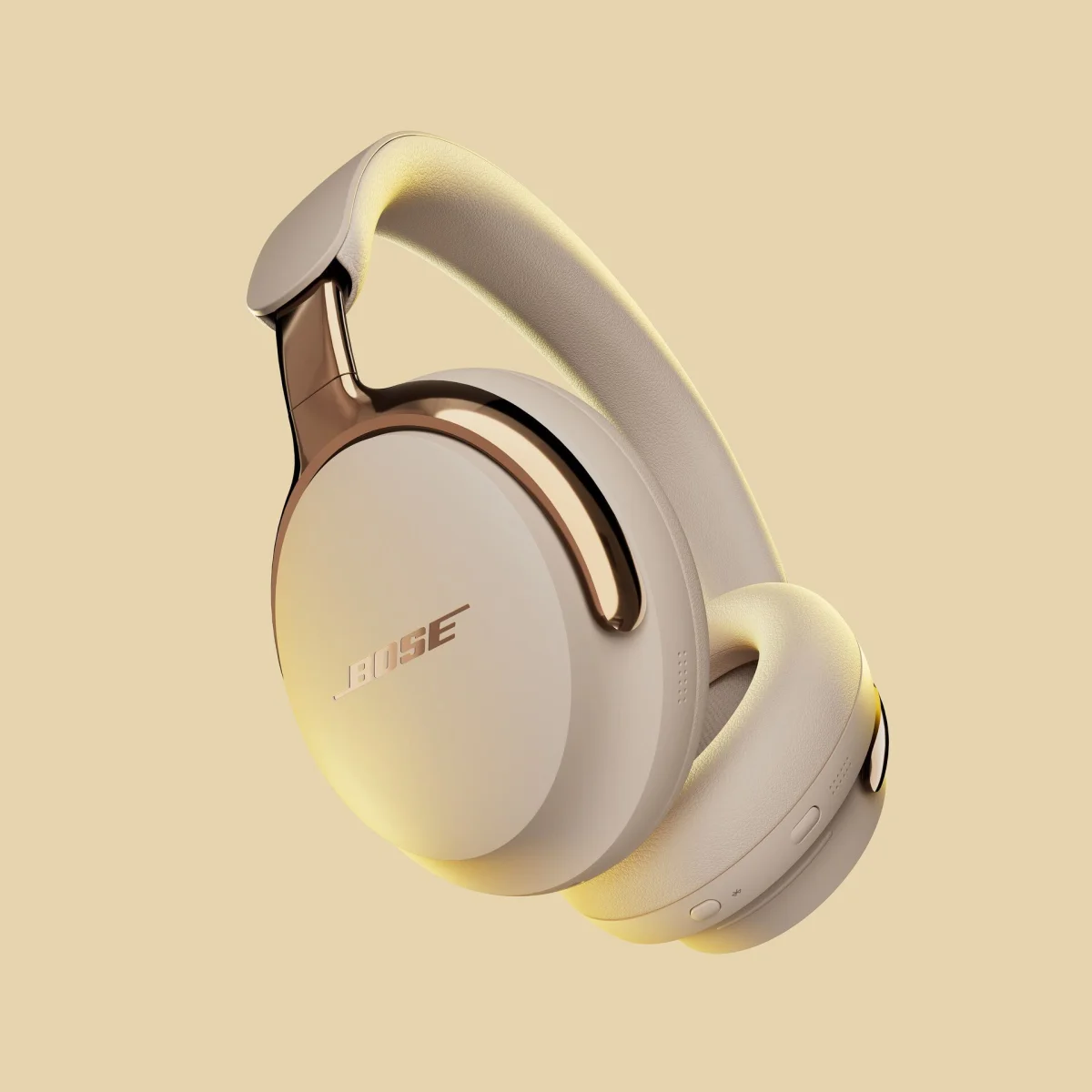 Over-ear Bose headphones in a beige color with rose gold accents on the ear cup hinges and the Bose logo. The headphones have cushioned ear pads and a padded headband, with control buttons visible on the bottom edge of one ear cup. The background is a matching beige color.