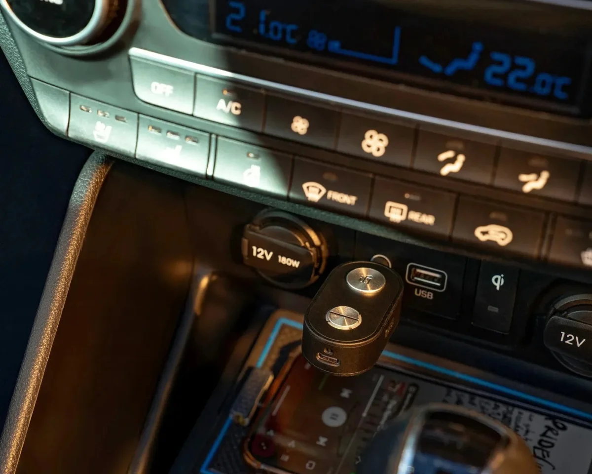 Compact, black dual-button car charger plugged into a 12V vehicle power outlet on the center console.
