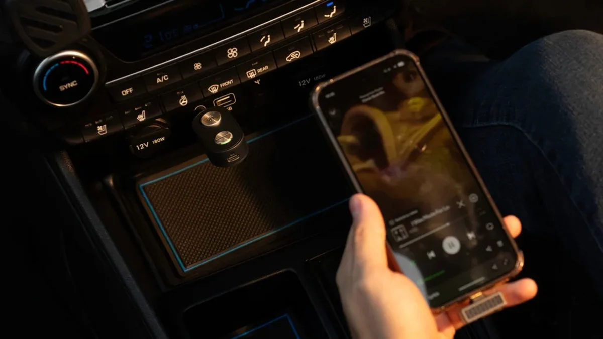 A smartphone held in a hand near a car center console with a Bluetooth adapter plugged into a 12V power outlet.