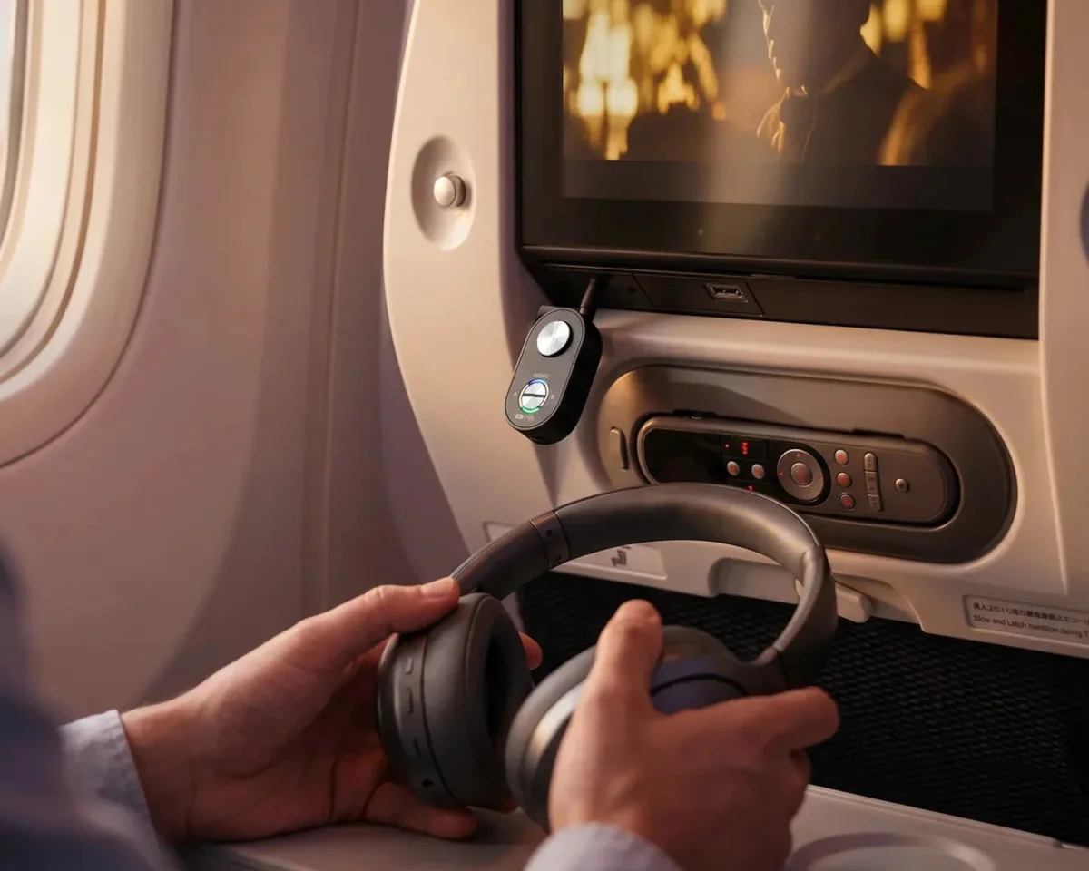 Wireless headphones held in hands in front of an airplane seatback screen and media control panel.