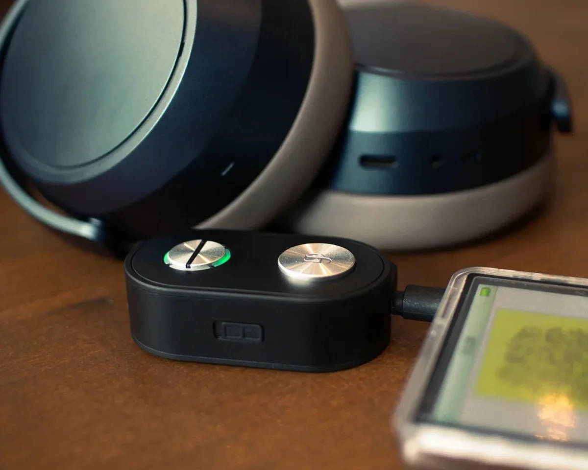 Black wireless headphones paired with a small, rectangular Bluetooth transmitter connected to a smartphone.