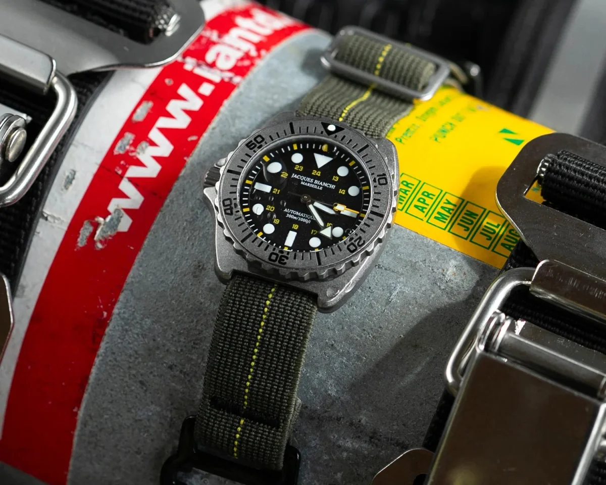 Diver's watch with black dial, luminous markers, and green fabric strap with yellow stitching on metal surface with red and yellow labels.
