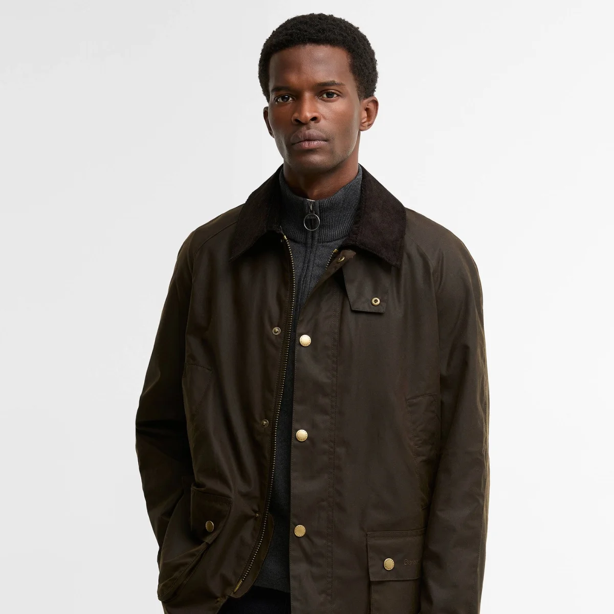 Dark olive waxed cotton jacket with a corduroy collar and gold-tone snap buttons, layered over a gray zip-up sweater.