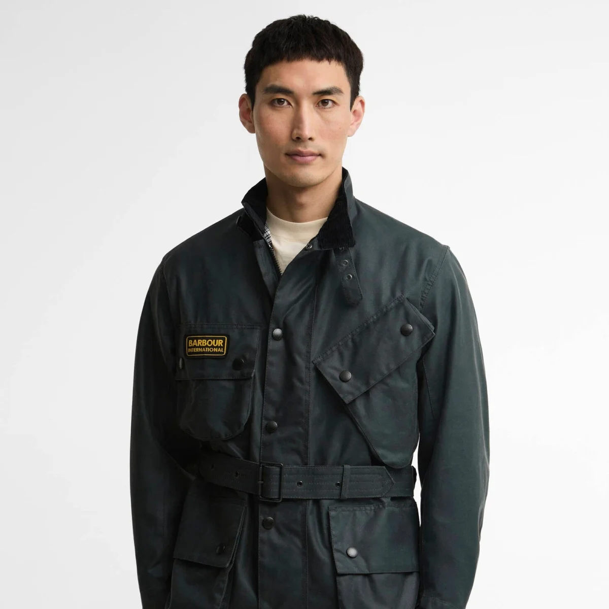 Dark green Barbour International waxed jacket featuring a belted waist, high collar, and a yellow brand logo patch on the chest.