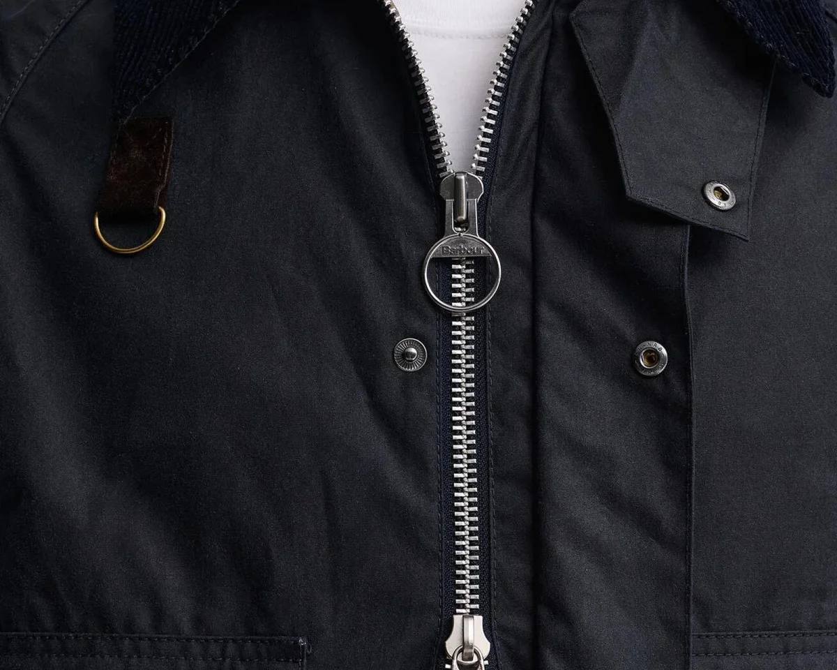 Close-up of a navy blue Barbour waxed jacket, featuring a corduroy collar, metal snap buttons, and a metal zipper pull with the brand logo.