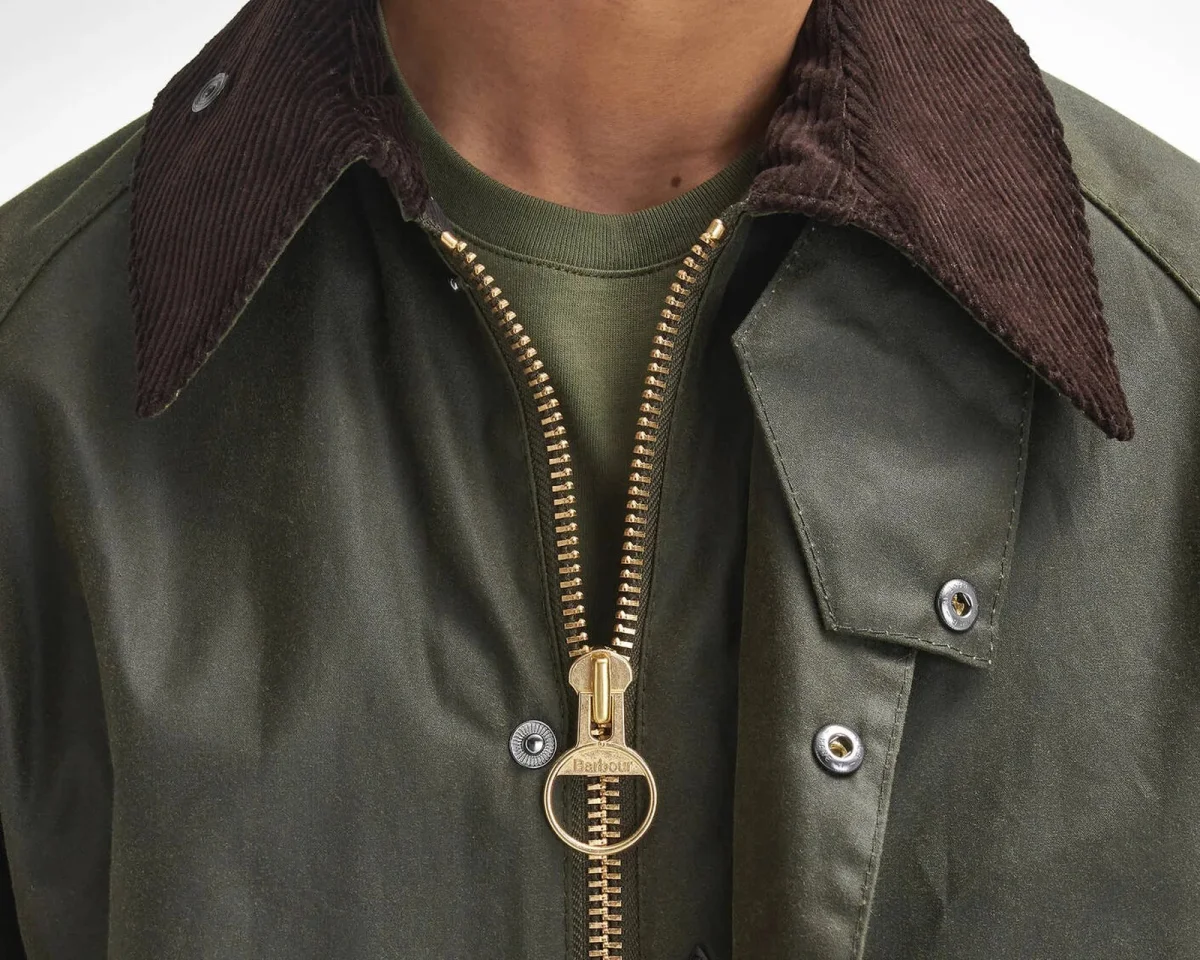 Olive green waxed jacket with a dark brown corduroy collar and a gold-toned circular zipper pull featuring the Barbour logo.