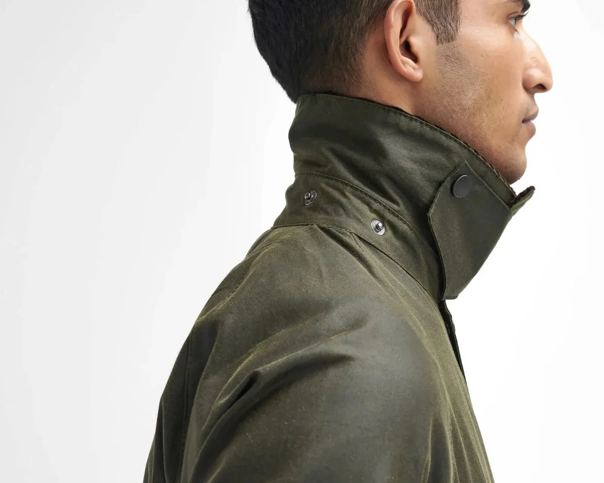 Profile view of an olive green waxed cotton jacket with a high collar and snap button closure.