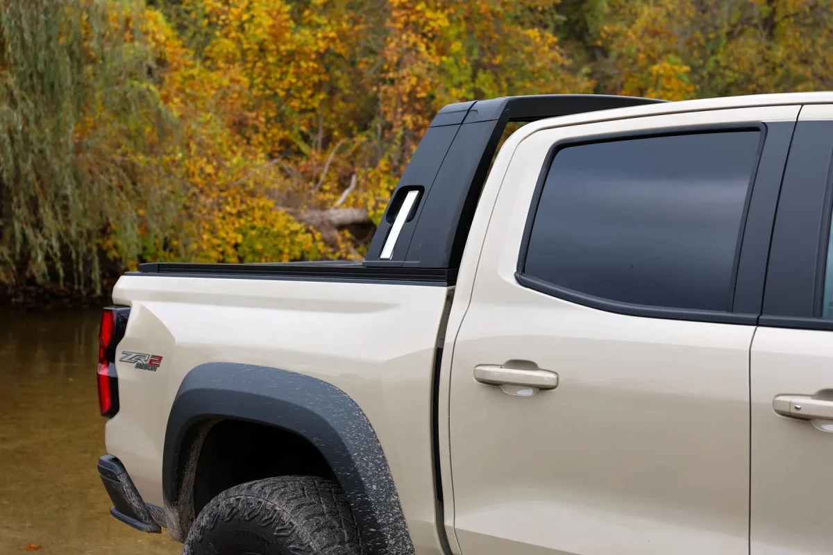 Tan crew cab pickup truck with a black sport bar in the truck bed, parked outdoors near trees with autumn foliage.