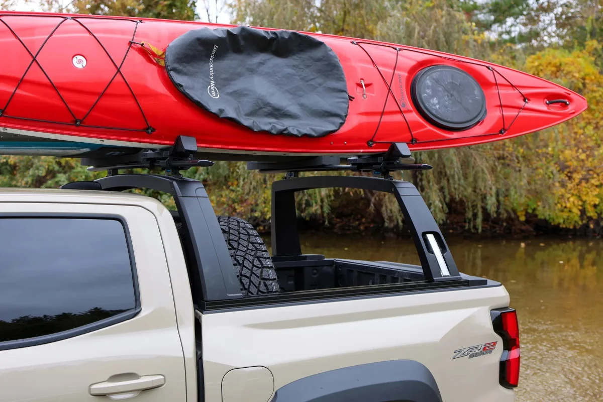 Red kayak with a black cockpit cover, secured to the bed rack of a tan pickup truck.
