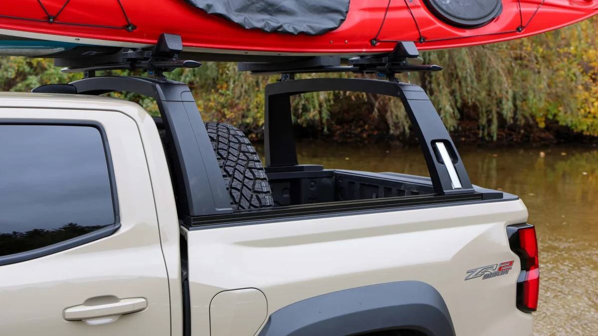 Tan pickup truck featuring a black bed rack holding a red kayak, parked near a body of water.
