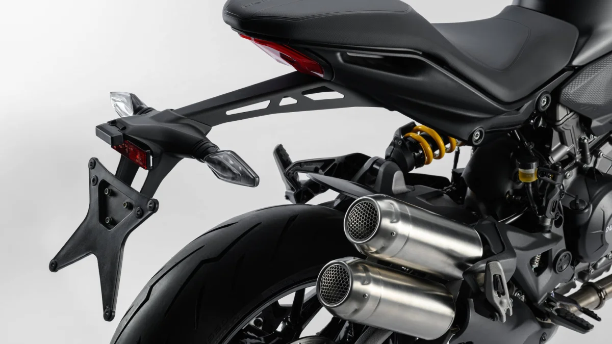Rear view of a motorcycle featuring a black seat, double metallic exhaust pipes with hexagonal mesh covers, a yellow rear suspension spring, and a license plate holder.