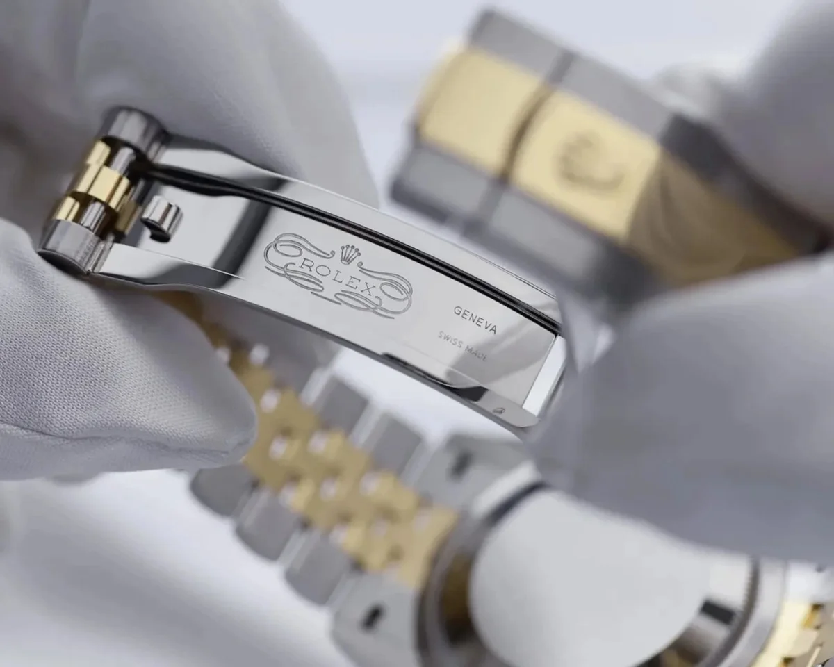Two-tone Rolex watch bracelet held in a gloved hand, showing the open internal clasp engraved with the Rolex logo, Geneva, and Swiss Made.