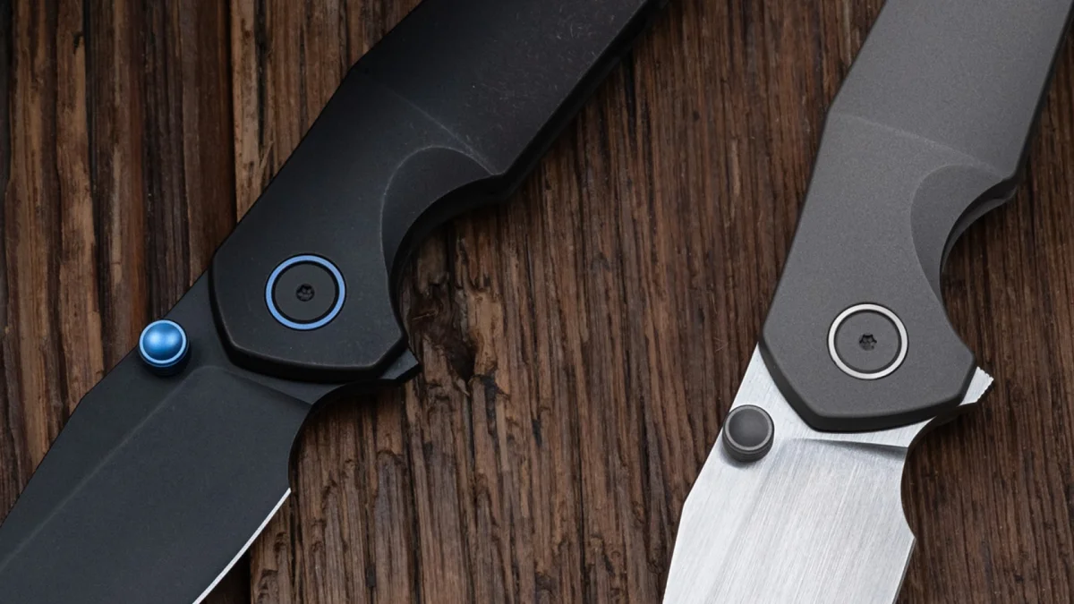 Two folding knives resting on a wooden surface; one featuring a black blade with blue accents, and one featuring a metallic blade with a grey finish.