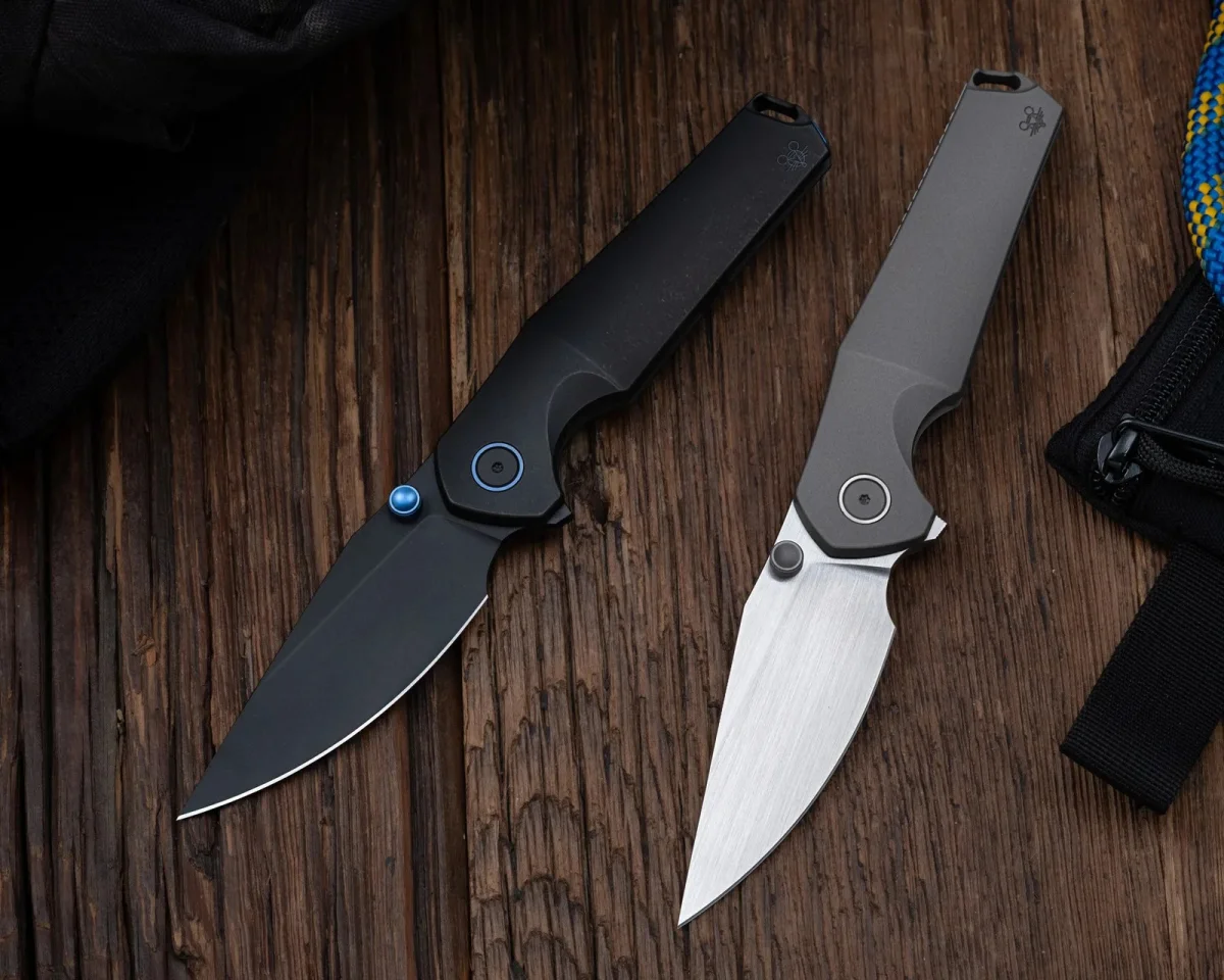 Two folding pocket knives with metal handles, one matte black with blue accents and one grey with a brushed silver blade, resting on a dark wood surface.